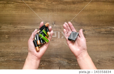 Top view of palms holding many disposable batteries and one rechargeable battery on wooden table, copy space. Concept of smart choice, comparison, saving money, reusable power, waste reduction 143358834