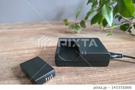 Rechargeable battery lying beside charger with cable on wooden table near green plant, copy space. Concept of reusable power, energy saving, eco friendly technology, smart choice, sustainability 143358866