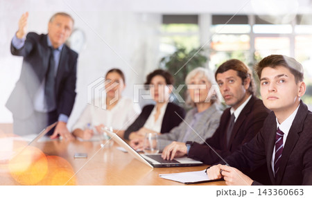 Office coworkers of different ages watching presentation while sitting at table with laptop and notes Office coworkers of different ages watching presentation while sitting at table with laptop and notes 143360663