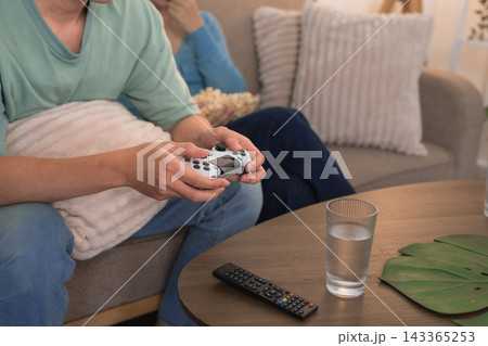 Asian couple relaxing on sofa playing video game together with popcorn and water glass nearby Asian couple relaxing on sofa playing video game together with popcorn and water glass nearby 143365253