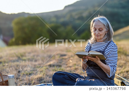Older woman enjoying peaceful solo picnic and reading in countryside. Older woman enjoying peaceful solo picnic and reading in countryside. 143367798