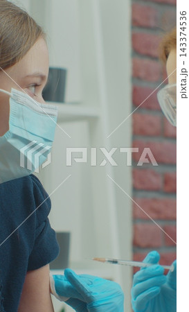 Vertical video. Female doctor wearing face mask injects a vaccine into the girl shoulder at home sitting on a couch. self isolation medical communication at home during pandemic. Vertical video. Female doctor wearing face mask injects a vaccine into the girl shoulder at home sitting on a couch. self isolation medical communication at home during pandemic. 143374536