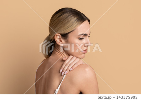 Young woman touching her shoulder and looking uncomfortable against beige studio background Young woman touching her shoulder and looking uncomfortable against beige studio background 143375905