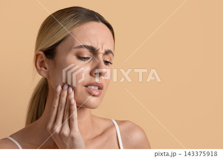Young woman holding her jaw in discomfort against beige studio background 143375918