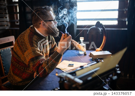 Pensive writer sits behind his desk and holds a smoking pipe to his lips with one hand Pensive writer sits behind his desk and holds a smoking pipe to his lips with one hand 143376057