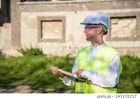 Construction professional using a tablet beside an unfinished building while wearing a hard hat and safety vest. Construction technology digital documentation and field reporting Construction professional using a tablet beside an unfinished building while wearing a hard hat and safety vest. Construction technology digital documentation and field reporting 143378157