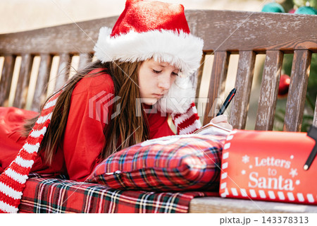 Christmas in July with girl wearing Santa hat writing letter to Santa outdoors beside decorated Christmas tree, Santa mailbox and festive decorations in sunny summer garden. 143378313