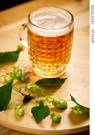 Large pint of beer with foam and sprigs of hops on wooden table 143379300