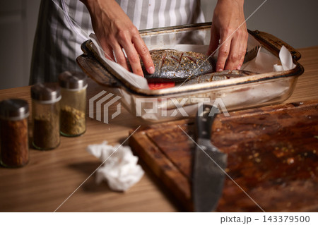 Chef hands seasoning whole sea bream fish in a baking dish, preparing a healthy seafood meal in the kitchen 143379500