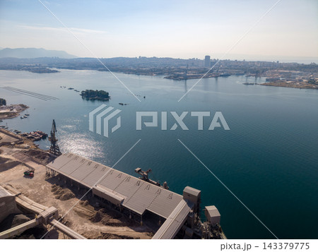 Industrial harbor sits beside sprawling coastal city, with loading crane, aggregate piles, and long covered storage sheds along water. Small wooded island and hazy mountain ridge mark bay. Industrial harbor sits beside sprawling coastal city, with loading crane, aggregate piles, and long covered storage sheds along water. Small wooded island and hazy mountain ridge mark bay. 143379775