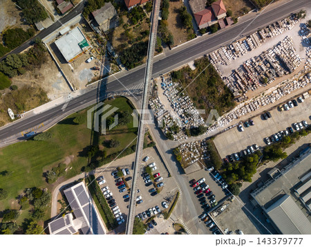 Overhead view of elevated rail line crossing busy roadway near stone depot. Railway bridge spans traffic lanes beside rows of stored blocks, parking areas, and patches of greenery. 143379777
