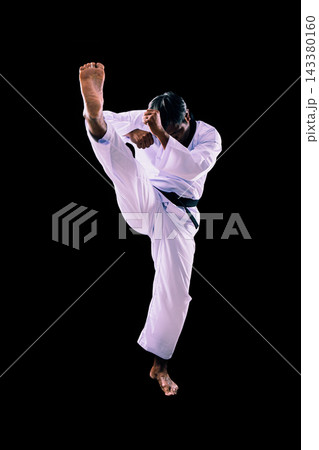 Martial artist performing powerful high kick in white uniform with black belt on dark background, showing karate, combat, strength, balance, discipline and athletic motion 143380160
