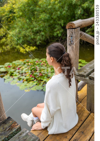 Young woman with dark hair wearing a white sweater sits on wooden dock beside a pond with lily pads and pink flowers in a serene natural setting Young woman with dark hair wearing a white sweater sits on wooden dock beside a pond with lily pads and pink flowers in a serene natural setting 143380559