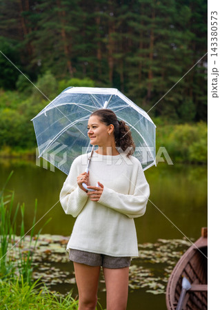 Young woman with curly hair holding a transparent umbrella stands by a serene pond surrounded by lush greenery and a wooden boat in the background Young woman with curly hair holding a transparent umbrella stands by a serene pond surrounded by lush greenery and a wooden boat in the background 143380573