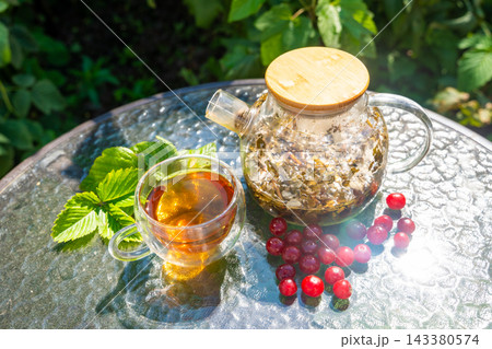 Glass cup and teapot with herbal tea served with fresh cherries and garden leaves. Homemade infusion represents preparing drinks from garden plants and berries Glass cup and teapot with herbal tea served with fresh cherries and garden leaves. Homemade infusion represents preparing drinks from garden plants and berries 143380574