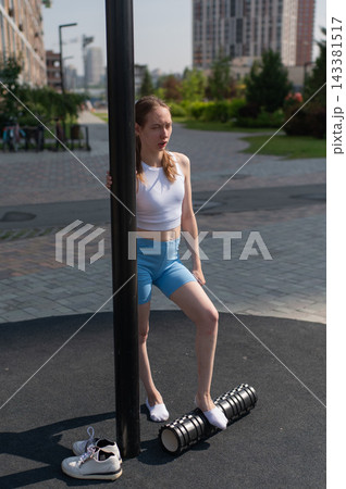 A Caucasian woman stretches her feet with a foam roller at a sports ground. A Caucasian woman stretches her feet with a foam roller at a sports ground. 143381517