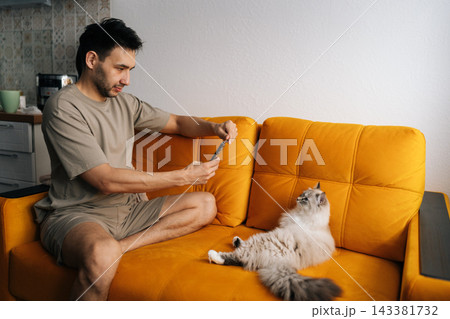 Happy young man sitting on bright orange sofa photographing fluffy cat with smartphone. Relaxed domestic lifestyle scene showing human animal bond and social media content creation at home. 143381732