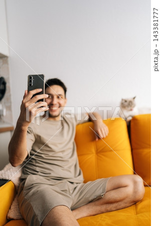 Cheerful young man in casual clothes sits on bright yellow couch using smartphone for selfie or video call. Pet cat rests in background. Relaxed home lifestyle scene with copy space. Natural lighting. Cheerful young man in casual clothes sits on bright yellow couch using smartphone for selfie or video call. Pet cat rests in background. Relaxed home lifestyle scene with copy space. Natural lighting. 143381777