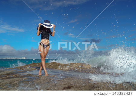 Woman in wide brim hat on rocky shore with splashing ocean wave Woman in wide brim hat on rocky shore with splashing ocean wave 143382427