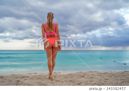 Woman in bright swimsuit standing on beach facing turquoise sea Woman in bright swimsuit standing on beach facing turquoise sea 143382457