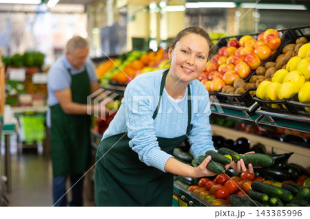 Middle-aged saleswoman putting cucumbers on food stall in greengrocery 143385996