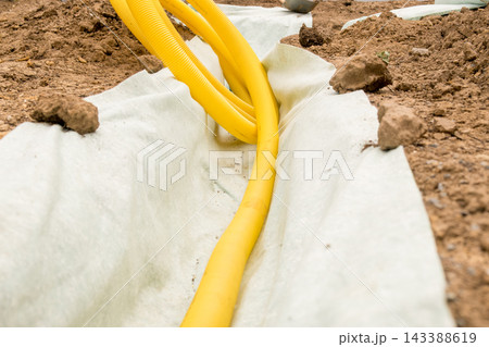 Within a landscaping trench, protective fabric supports a yellow drainage hose arranged for effective water management. The setup emphasizes an organized installation process. Within a landscaping trench, protective fabric supports a yellow drainage hose arranged for effective water management. The setup emphasizes an organized installation process. 143388619