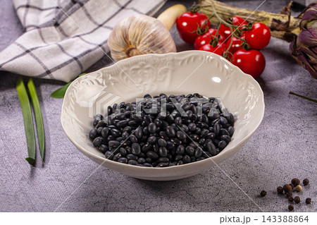 Black beans in ornate white ceramic bowl surrounded by cherry tomatoes garlic green onions and peppercorns on textured grey surface 143388864