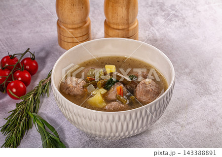 White ceramic bowl filled with meatball soup containing potatoes green beans carrots and herbs on textured grey surface with cherry tomatoes and rosemary sprig nearby food dish White ceramic bowl filled with meatball soup containing potatoes green beans carrots and herbs on textured grey surface with cherry tomatoes and rosemary sprig nearby food dish 143388891