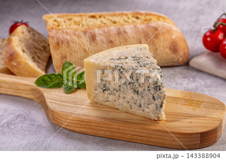 Wooden cutting board with wedge of blue cheese and fresh basil leaves next to rustic bread loaf and red cherry tomatoes on grey stone surface appetizer arrangement Wooden cutting board with wedge of blue cheese and fresh basil leaves next to rustic bread loaf and red cherry tomatoes on grey stone surface appetizer arrangement 143388904