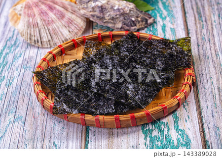 Dried nori seaweed sheets on round woven bamboo tray with red bindings and scallop shell on distressed teal wood surface Dried nori seaweed sheets on round woven bamboo tray with red bindings and scallop shell on distressed teal wood surface 143389028