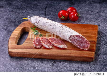 Cured sausage fuet on wooden cutting board with sliced pieces and cherry tomatoes beside it, food preparation Cured sausage fuet on wooden cutting board with sliced pieces and cherry tomatoes beside it, food preparation 143389061