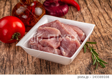 Rustic wooden table surface with raw pork chunks in white square bowl surrounded by fresh tomato and rosemary ingredient preparation 143389108