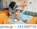 Young woman sitting on a bed while changing a bandage on her injured leg at home. Medical supplies, gauze, scissors, and home care items illustrate wound treatment, first aid, rehabilitation, and 143389738