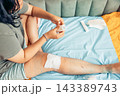 plus size young woman sitting on bed and applying a gauze bandage to her injured leg at home. visible scars and bruising on the knee and thigh. home first aid, wound care, injury recovery 143389743
