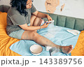 plus size young woman sitting on a bed and cutting gauze for dressing an injured leg at home. visible scars and healing wounds on her knee and lower leg. home first aid, wound care, injury recovery 143389756