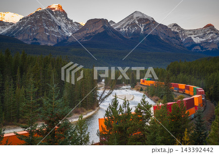 Serene freight train travels through mountain forest along river in Canada showcasing peaceful wilderness scenery during sunset 143392442