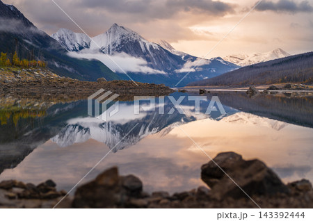 Serene mountain lake reflection reveals peaceful nature under golden sunset in Canada with snowy peak mirroring calm water Serene mountain lake reflection reveals peaceful nature under golden sunset in Canada with snowy peak mirroring calm water 143392444