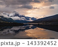 Serene canada mountain wilderness offering peaceful reflection on calm lake water beneath snow peak landscape during dramatic golden sunset 143392452