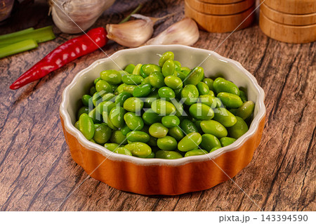 Wooden table with ceramic bowl holding bright green edamame beans, red chili pepper, garlic cloves, and green onions nearby, food presentation 143394590