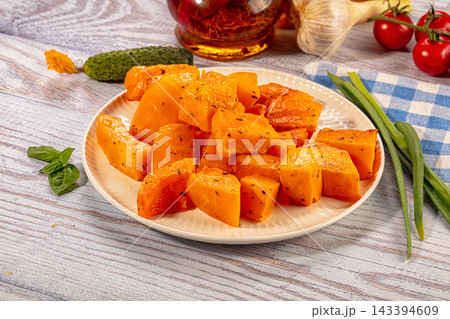 Baked pumpkin pieces seasoned with herbs on white plate with garlic and tomatoes on wooden table 143394609