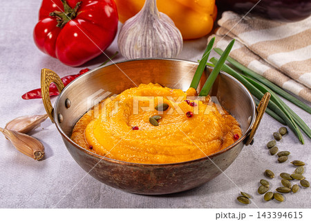 Metal bowl with ornate handles holding creamy orange pumpkin soup garnished with green pumpkin seeds and red peppercorns, surrounded by fresh vegetables and herbs 143394615