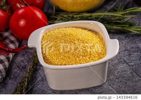 Dark stone surface with white ceramic bowl filled with raw yellow millet grains and fresh red tomatoes, rosemary sprigs, dried chili pepper, checkered cloth, healthy organic cereal ingredient 143394616