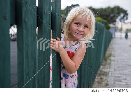 Cute Little Blonde Girl Peeking Behind Green Wooden Fence Outdoors 143396508