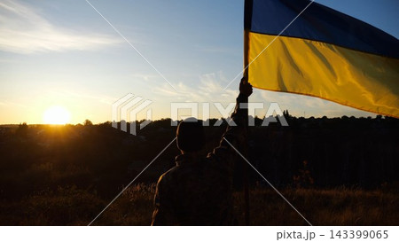 Male military in uniform lifting blue-yellow banner in honor of the victory against russian aggression at sunset. Young soldier of ukrainian army raised a waving flag of Ukraine at countryside 143399065