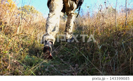 Low view to male feet of ukrainian soldiers going along trail at countryside. Legs of young army man walking through dry grass at sunny day. War in Ukraine. Slow motion Low view to male feet of ukrainian soldiers going along trail at countryside. Legs of young army man walking through dry grass at sunny day. War in Ukraine. Slow motion 143399085