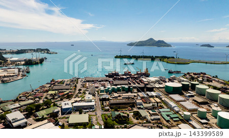 Harbor with industrial buildings, storage tanks, containers, and turquoise sea, with green islands in the background. Seychelles, Mahe. Victoria. 143399589