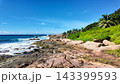 Rocky coastline with turquoise waves crashing against the shore, surrounded by lush green vegetation under a blue sky. Seychelles, La Digue. 143399593