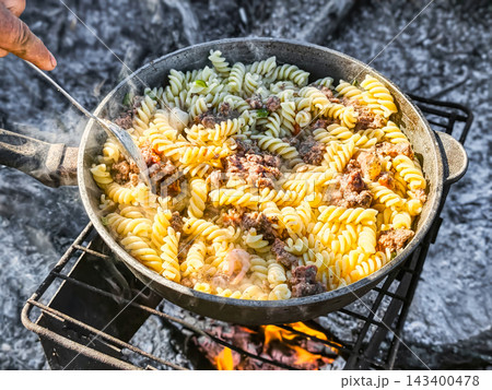 Pasta is being prepared with meat on a campfire at a nature site. Steam rises from the pot as food is mixed. This is a group activity enjoyed in the Pasta is being prepared with meat on a campfire at a nature site. Steam rises from the pot as food is mixed. This is a group activity enjoyed in the 143400478