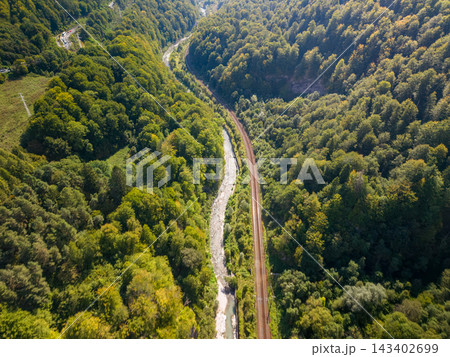 Railway follows river through forested gorge in Romania, with parallel tracks curving between wooded slopes and pale rocky stream. Aerial landscape highlights remote mountain rail access. Railway follows river through forested gorge in Romania, with parallel tracks curving between wooded slopes and pale rocky stream. Aerial landscape highlights remote mountain rail access. 143402699