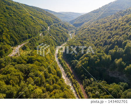 Romanian mountain valley extends between forested slopes, with river, railway and winding road visible below. Romanias wooded landscape forms broad aerial panorama in warm sunlight. Romanian mountain valley extends between forested slopes, with river, railway and winding road visible below. Romanias wooded landscape forms broad aerial panorama in warm sunlight. 143402701